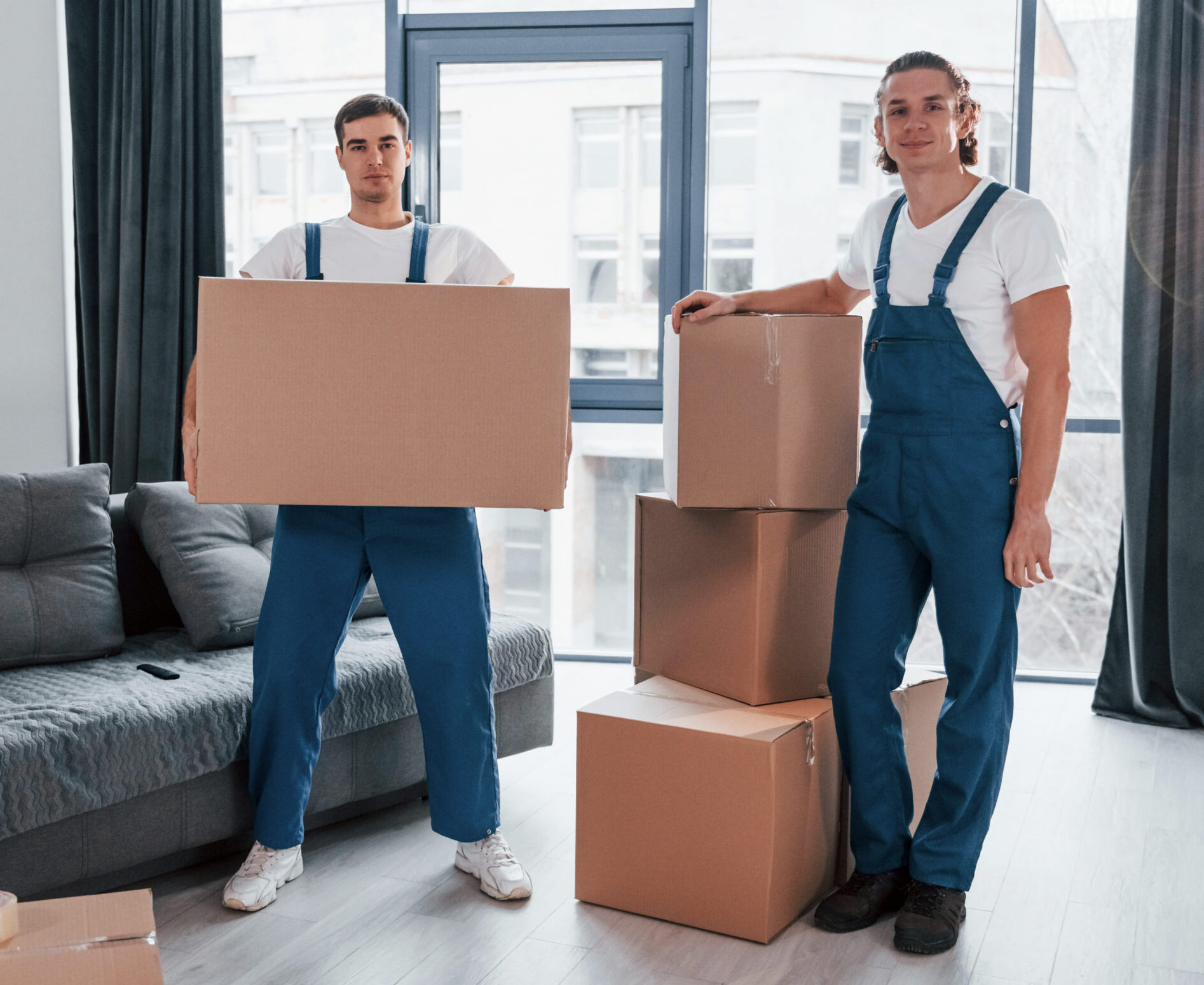 Busy day. Two young movers in blue uniform working indoors in the room